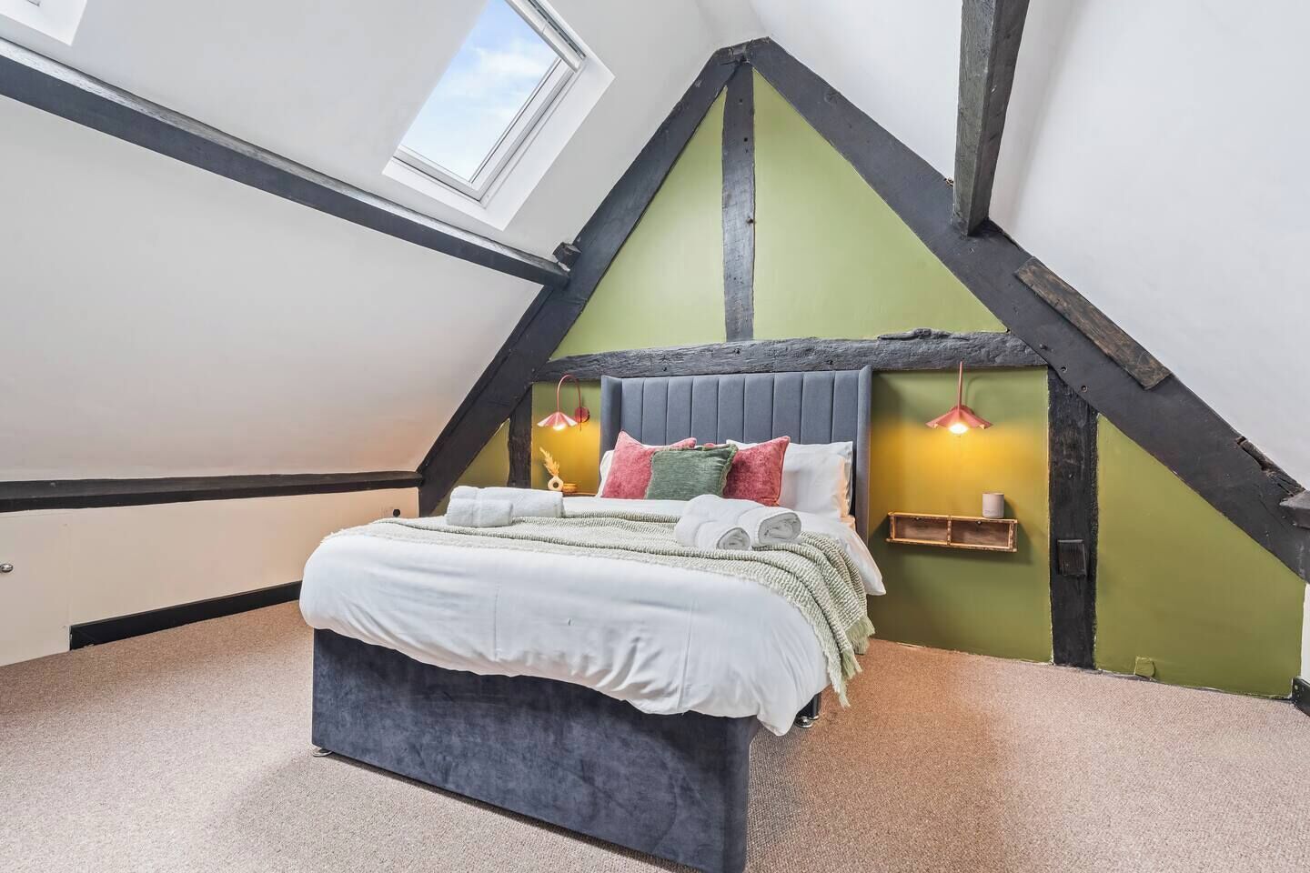 A bedroom with a sloped ceiling, a bed with multiple pillows, a bedside table with a lamp, and a window.