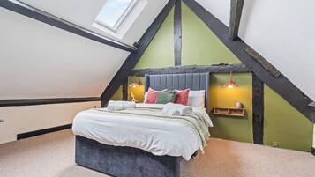 A bedroom with a sloped ceiling, a bed with multiple pillows, a bedside table with a lamp, and a window.