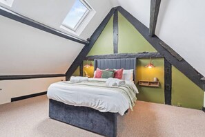 Ein Schlafzimmer mit schrägen Wänden, einem Bett mit vielen Kissen, einem Nachttisch mit Lampe und einem Fenster.