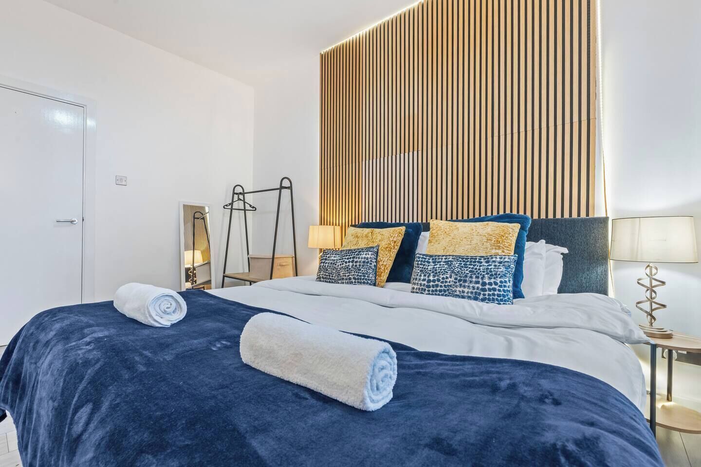 Una habitación de hotel moderna con una cama grande, un cabecero de listones de madera y una mesita de noche con una lámpara.