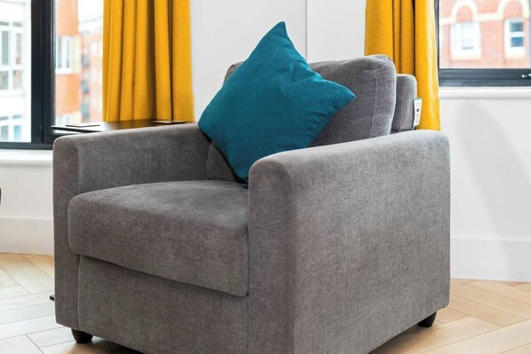 Un sillón gris con un cojín azul verdoso en una habitación con cortinas amarillas y vista al exterior.