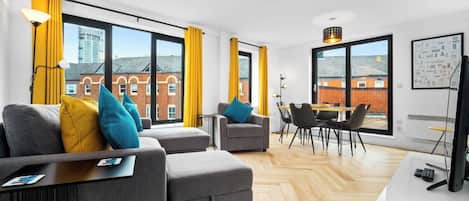 A modern living room with a sofa set, a dining area, and large windows with curtains.