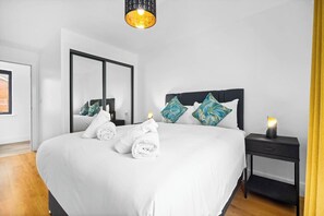 Un lit bien fait, avec du linge de lit blanc et des serviettes pliées, une table de chevet avec une lampe et un grand miroir au mur.