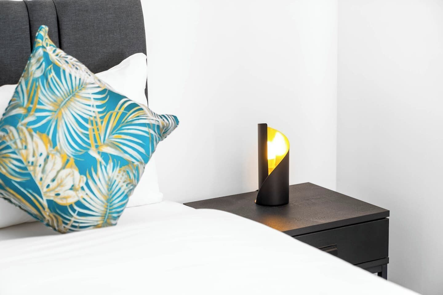 Ein Bett mit weißer Bettdecke und blau-weißem Kopfkissen, ein Nachttisch in Schwarz mit einer Lampe und ein grauer Kopfteil.