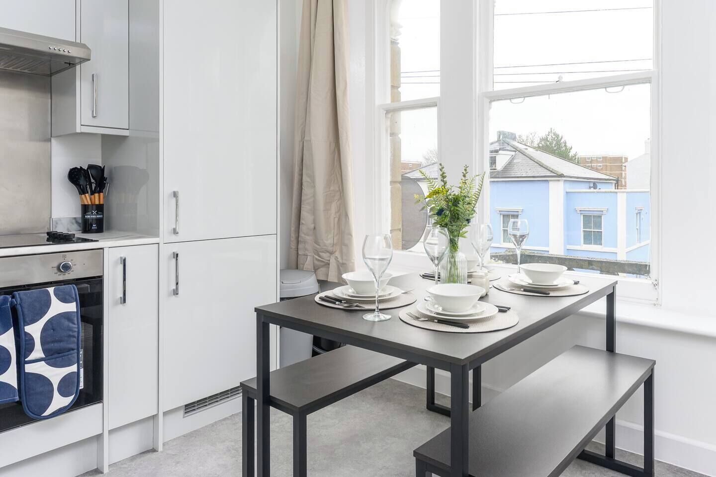 Een moderne keuken met witte kasten, een inbouwoven en een eettafel voor vier personen, gedekt met witte borden en glazen.