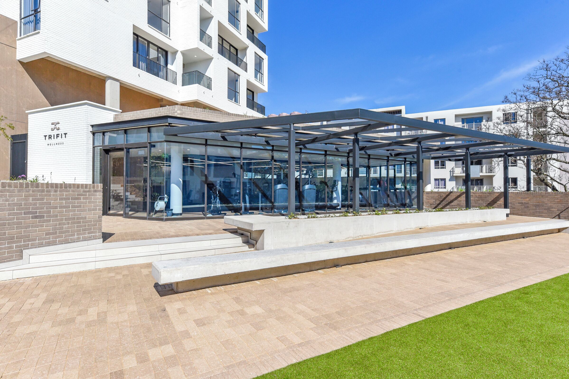 Un edificio moderno con fachada de vidrio, un área exterior cubierta y un letrero que dice 'TRIFIT WELLNESS'.