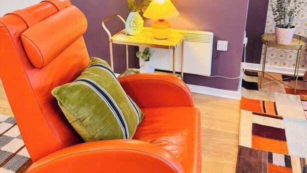 Ein Zimmer mit einem einzelnen orangefarbenen Sessel, einem runden Spiegel, einem kleinen Tisch mit Lampe und einer im Hintergrund sichtbaren Küche.