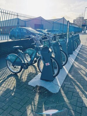 Eine Reihe von Fahrrädern des West Midlands Cycle Hire Systems, geparkt in einem überdachten Bereich mit einer Umzäunung und einem Gebäude im Hintergrund.