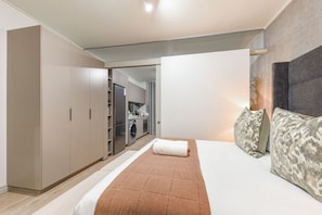 A bedroom with a bed, a wardrobe, a built-in washing machine, and a small kitchen area. - One Rosebank | Modern Comfort | Heart of the City (Johannesburg)