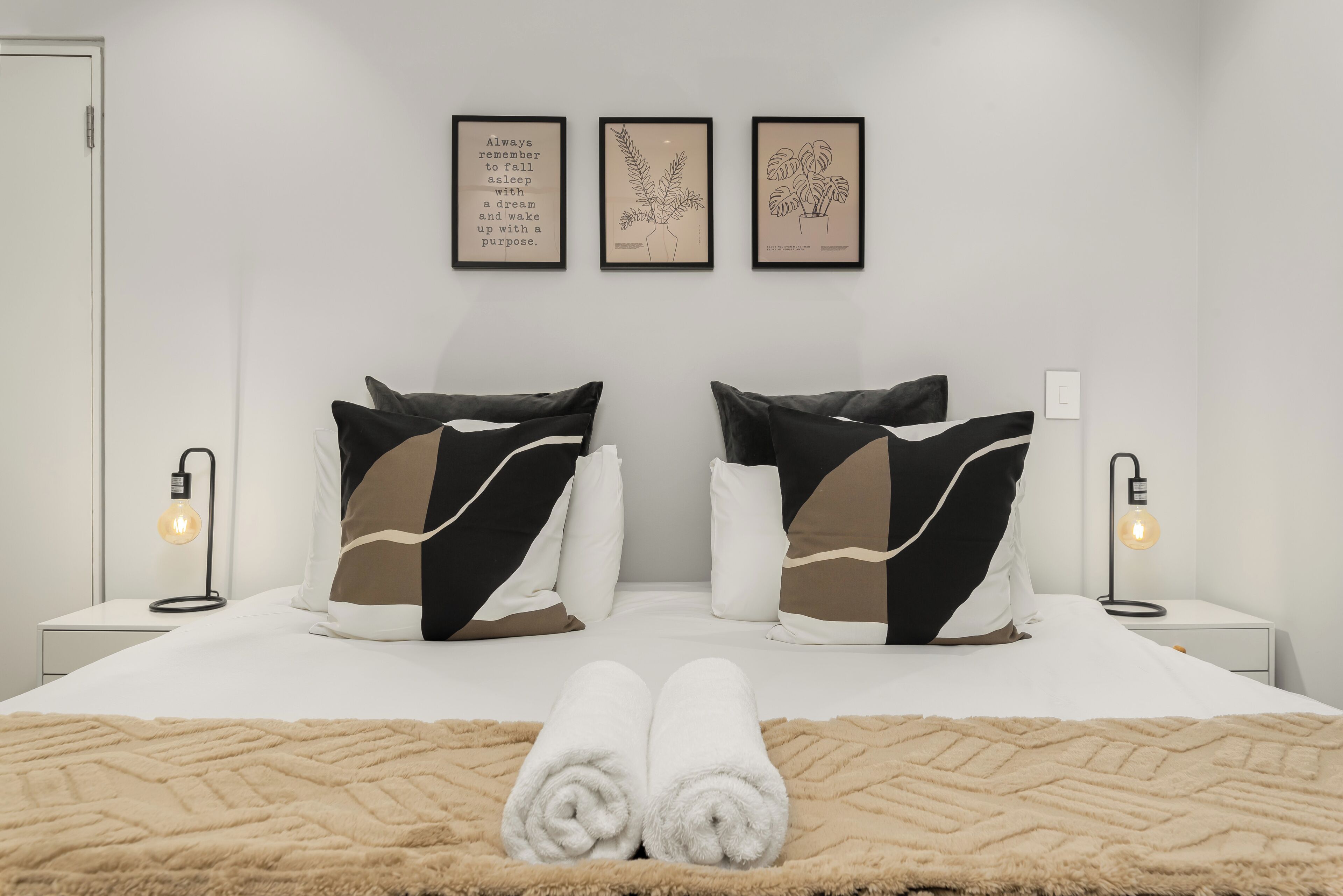 Ein ordentlich bezogenes Bett mit zwei Kissen, einem Paar zusammengerollter Handtücher und einer dekorativen Tagesdecke. Zwei Nachttischlampen und drei gerahmte Wandbilder befinden sich über dem Bett.
