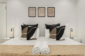 Un lit bien fait, avec deux oreillers, une pile de serviettes enroulées et un plaid à motifs. On y trouve deux lampes de chevet et trois œuvres d’art encadrées accrochées au mur au-dessus du lit.