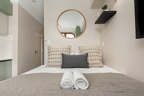 Un dormitorio con una cama, un espejo redondo, dos almohadas con textura, una toalla enrollada y una pequeña planta en maceta.