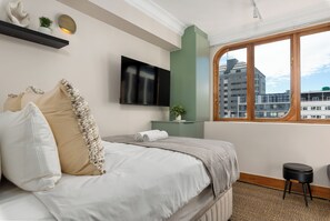 Ein modernes Schlafzimmer mit Bett, Flachbildfernseher, einem grünen Schrank, einem Fenster mit Blick auf Gebäude und einem kleinen Hocker.