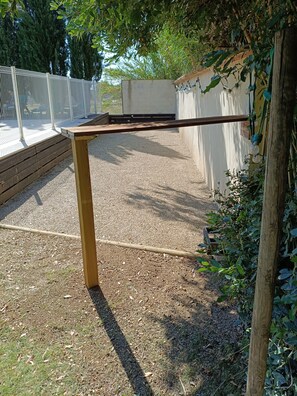Ein Holztisch mit Metallgestell steht auf einer Schotterfläche, im Hintergrund ist ein Holzzaun und viel Grün zu sehen.