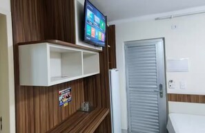Ein Hotelzimmer mit einem an einer Holzverkleidungung montierten Fernseher, einer weißen Tür und einem kleinen Regal mit einem Poster.