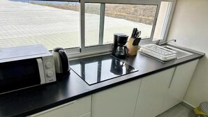 Une cuisine équipée d’un four à micro-ondes, d’une machine à café et d’ustensiles de cuisine, le tout posé sur le plan de travail, avec une fenêtre donnant sur une zone extérieure dont le sol est constitué d’un motif quadrillé.