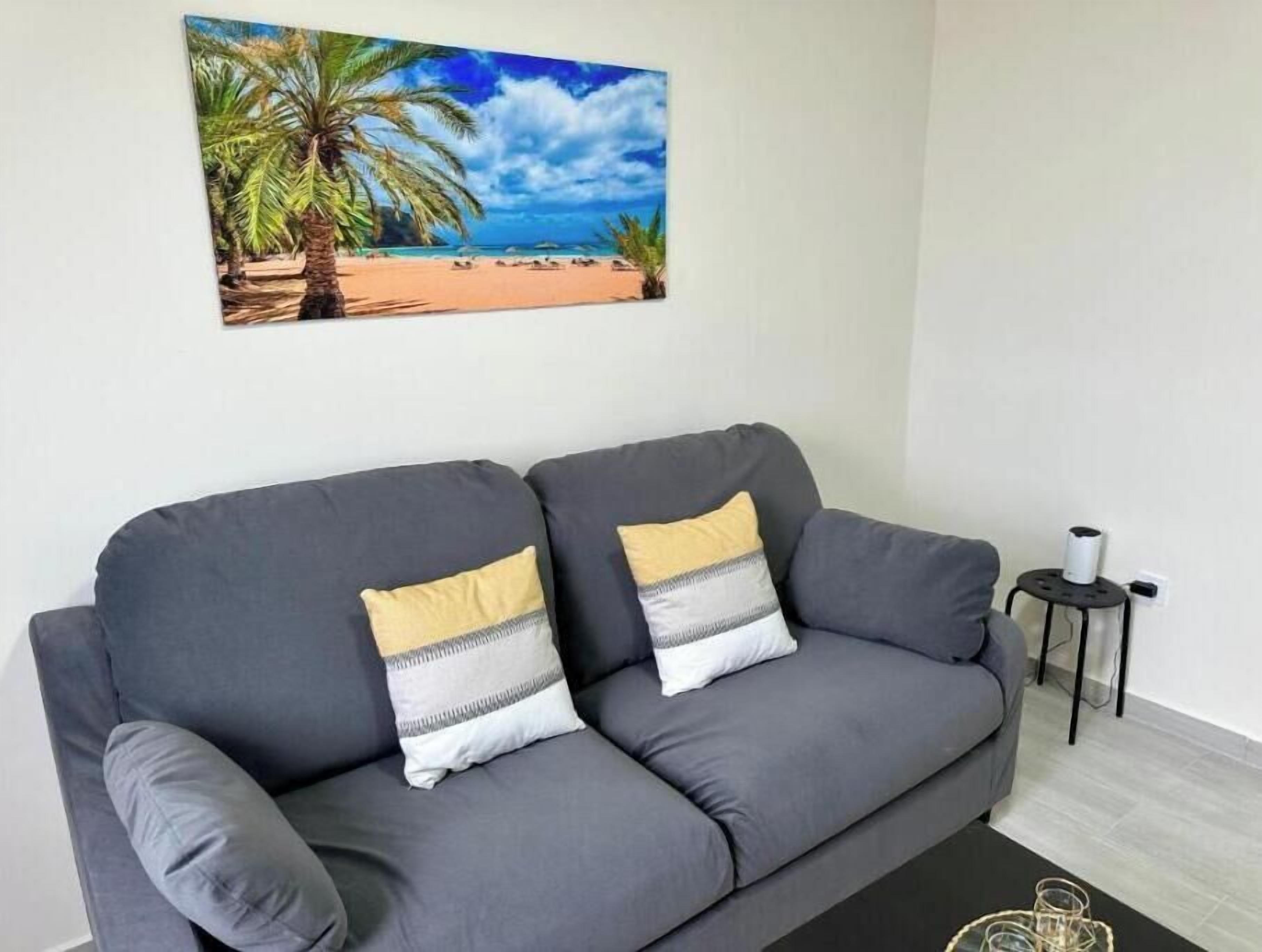 Een grijze bank met twee kussens, een klein zwart nachtkastje met een wit voorwerp, en een strandlandschap op de muur.