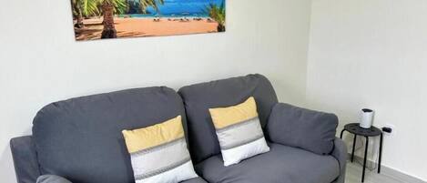 A grey sofa with two pillows, a small black side table with a white object, and a beach scene painting on the wall.