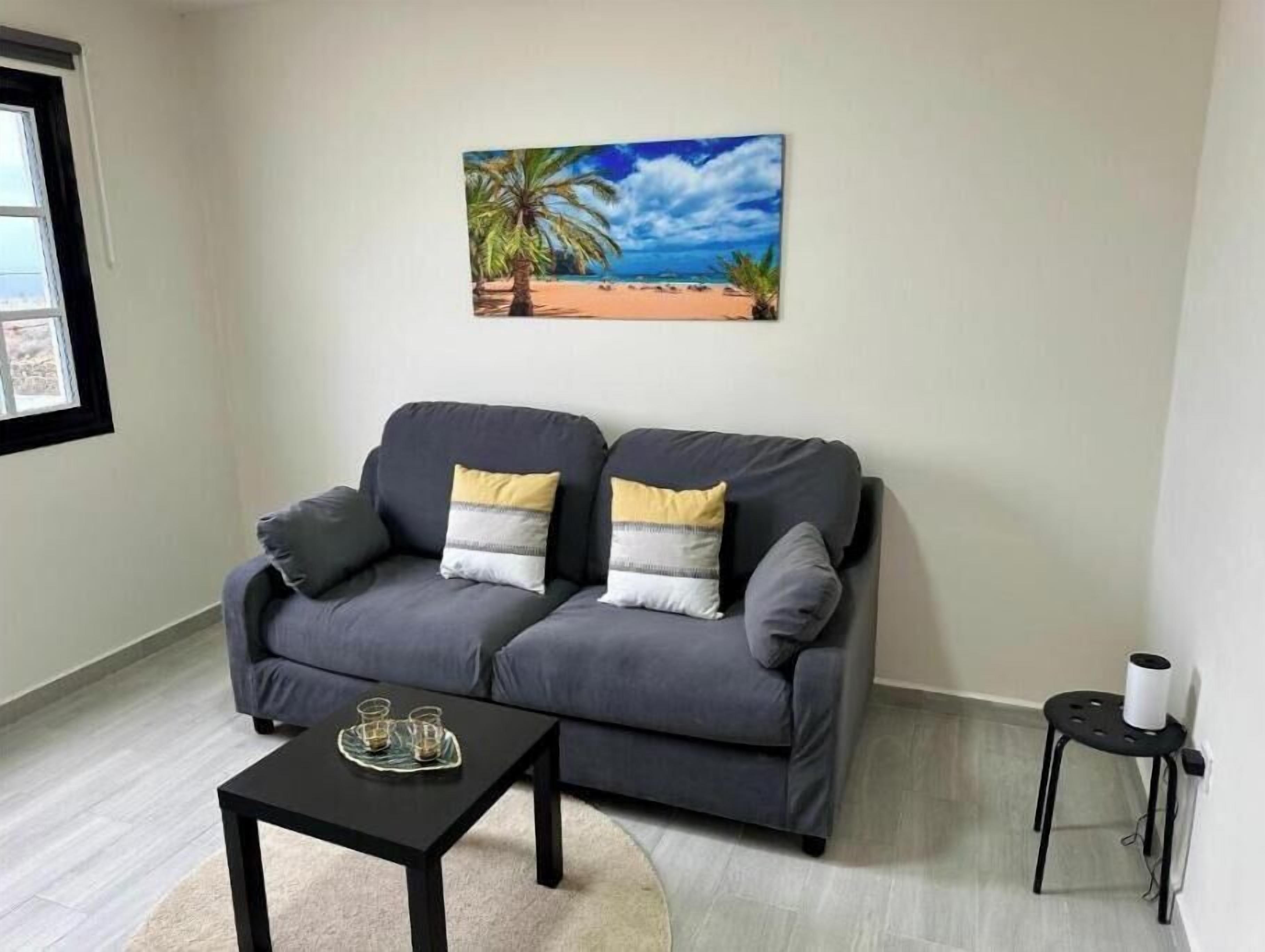Un canapé gris avec des coussins, une table basse noire, une petite table d’appoint noire sur laquelle se trouve un objet blanc, et un tableau représentant une scène de plage accroché au mur.