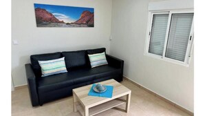 A living room with a black sofa, two pillows, a coffee table, a painting of a landscape, and a window with blinds. - New modern apartment, Scandinavian style (Icod de los Vinos)