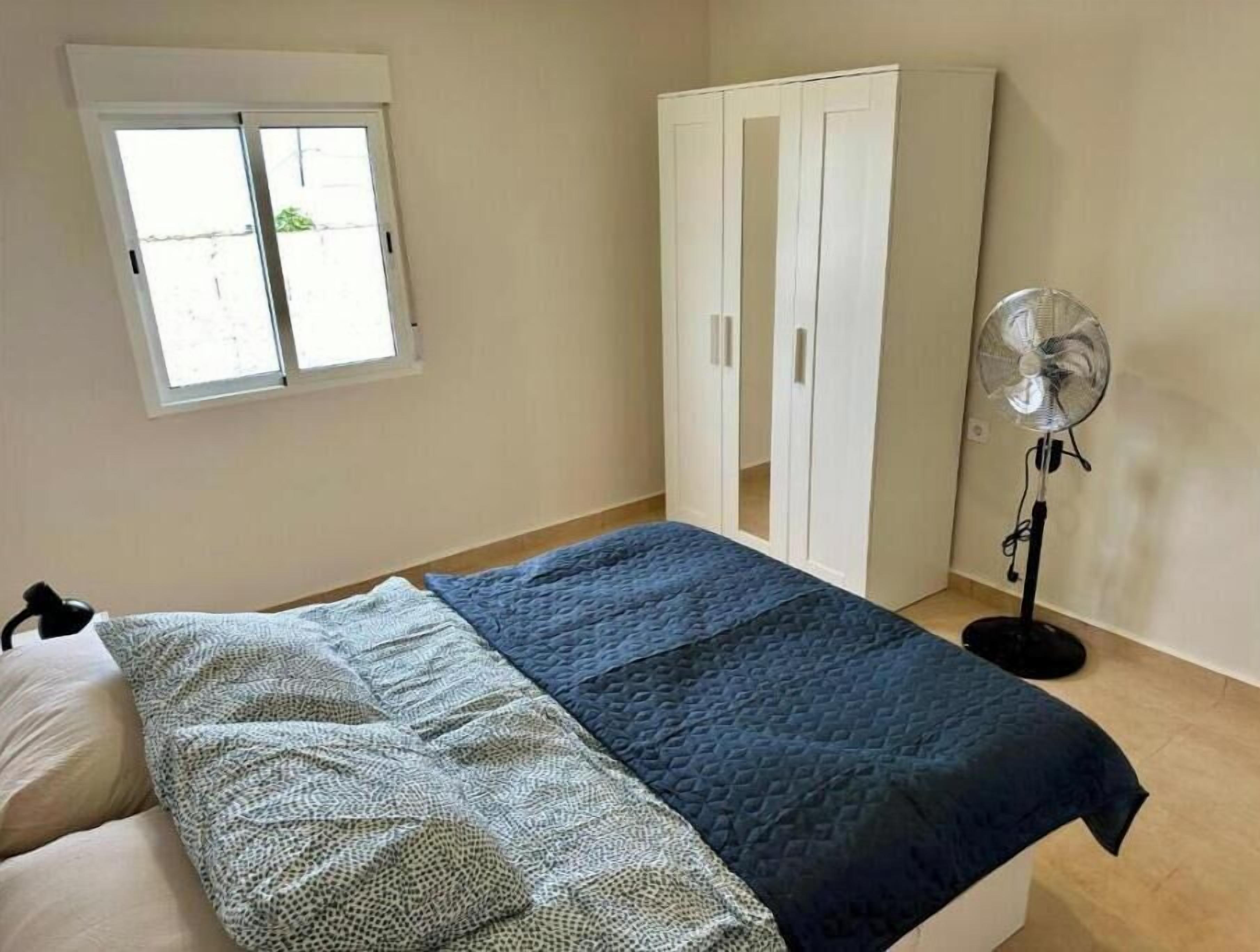 Une chambre à coucher avec un lit, une armoire, une fenêtre et un ventilateur sur pied.