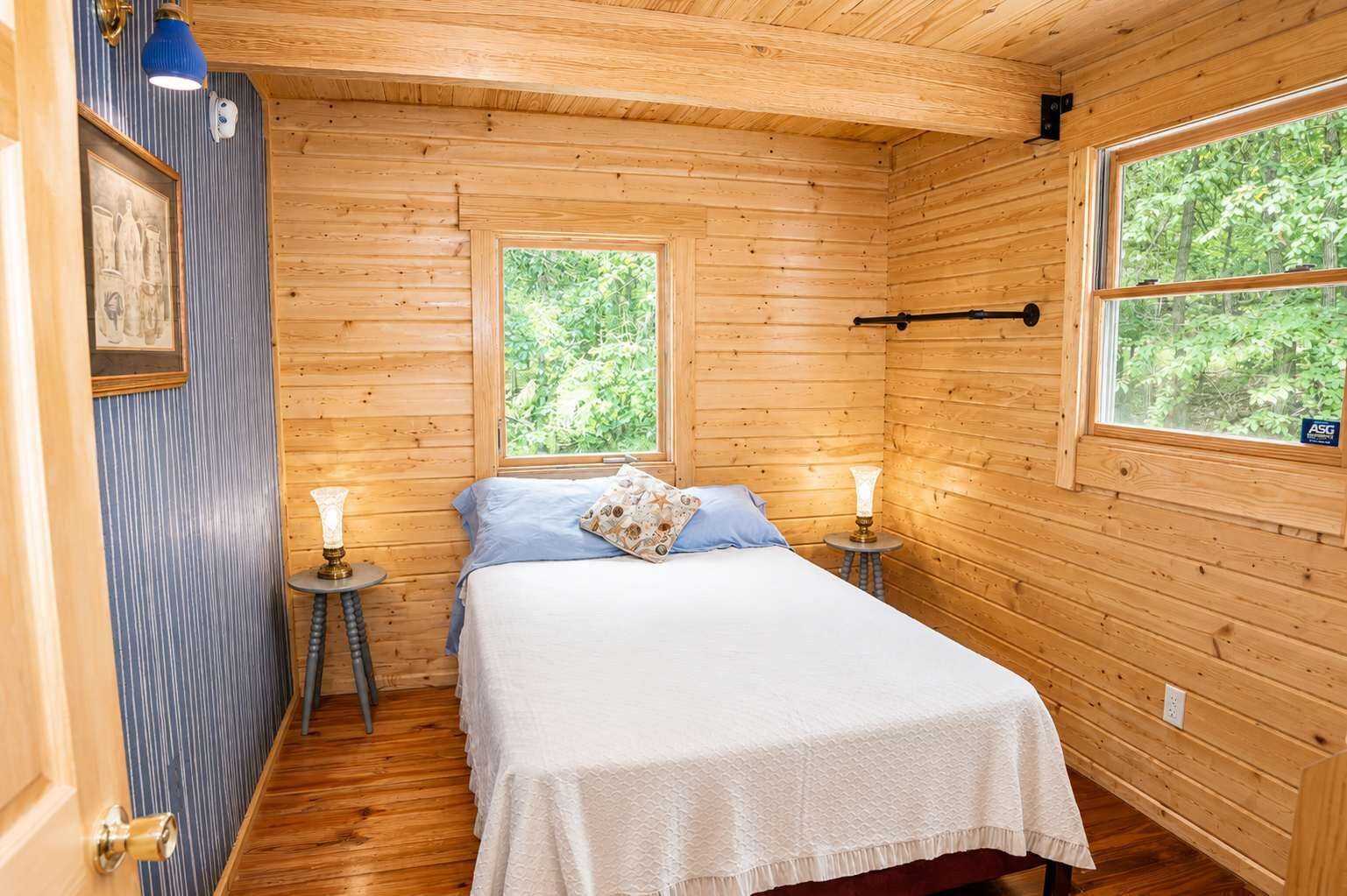 Una habitación de cabaña de madera con una cama, dos ventanas, una puerta y una mesita con una lámpara.