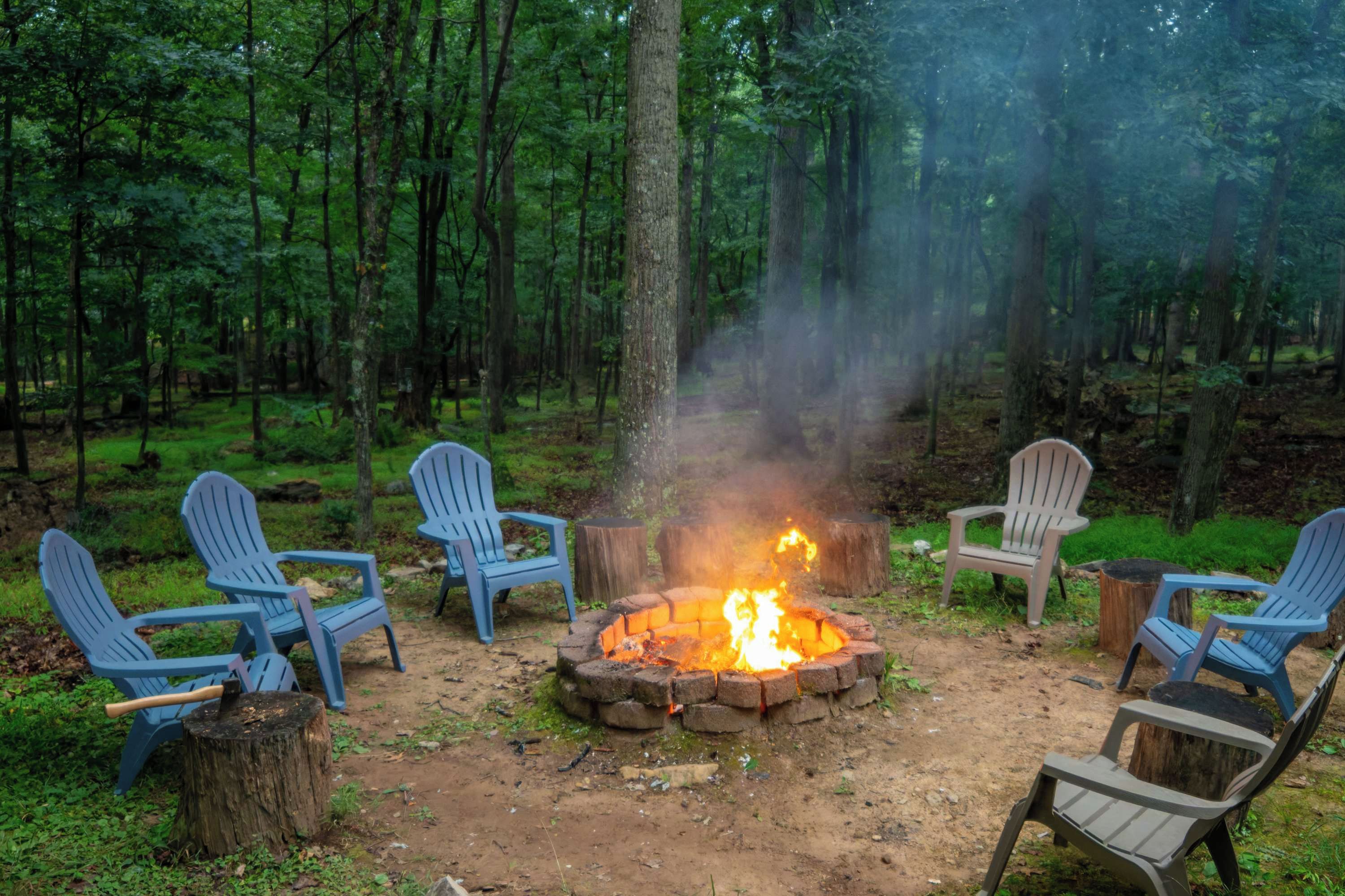 A campfire surrounded by four Adirondack chairs in a forest setting.