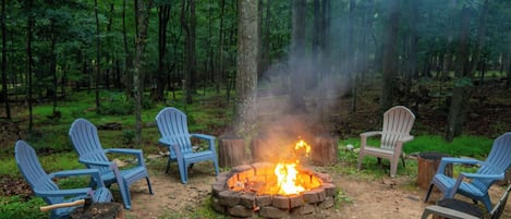 A campfire surrounded by four Adirondack chairs in a forest setting.