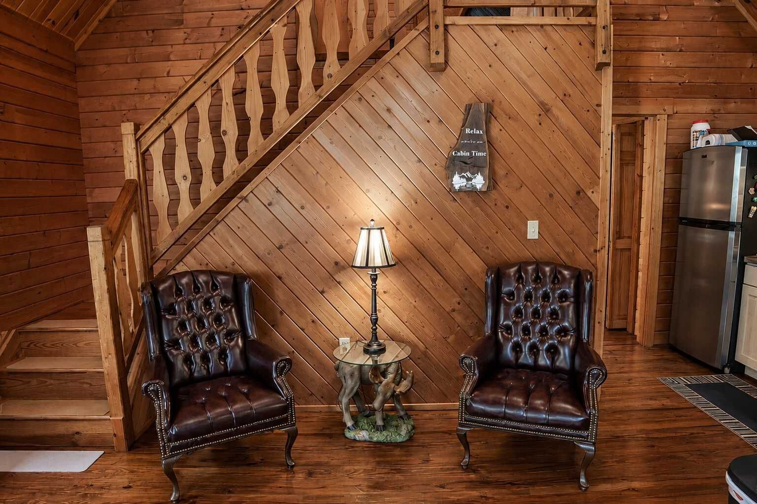 Interior de una cabaña con escalera de madera, dos sillones de cuero, una lámpara de mesa y un refrigerador.