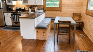 A compact kitchen with wooden cabinets, a stove, and a dining area with a bench and table. - River Cabin w/Kayaks, Fire Pit, WiFi & Game Room (Great Cacapon)
