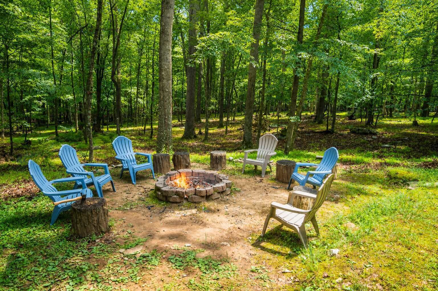A forest clearing with a fire pit and Adirondack chairs arranged around it.