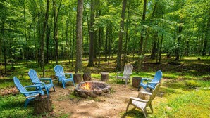 A forest clearing with a fire pit and Adirondack chairs arranged around it. - River Cabin w/Kayaks, Fire Pit, WiFi & Game Room (Great Cacapon)