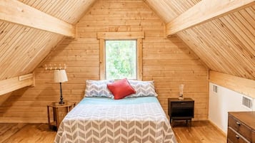 A bedroom with a wooden sloped ceiling, a bed with a patterned bedspread, a colorful rug, a nightstand, a dresser, and a window with a view of greenery.
