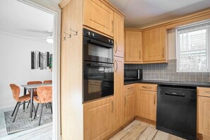 A modern kitchen with wooden cabinets, a black dishwasher, and a dining area with orange chairs. - #121 Two Beautiful cottages in Cornwall |for 14 (Cornwall)