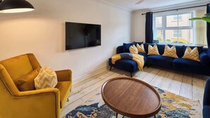 Een woonkamer met een bank, een fauteuil, een flatscreen televisie, een plafondventilator en een groot raam met gordijnen.