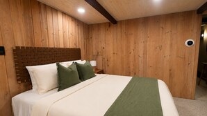 A bedroom with a wooden headboard, a bed with white and green bedding, a nightstand with a lamp, and a thermostat on the wall.