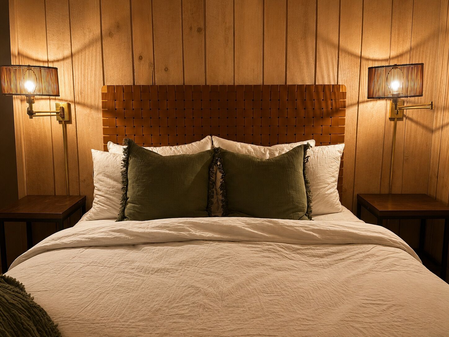 A hotel room with a wooden headboard, two bedside lamps, a bed with white linens, and two green pillows.