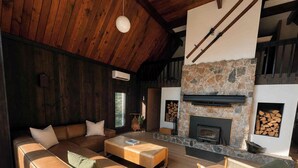 A cozy living room with a stone fireplace, wooden ceiling beams, and a large window.