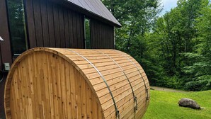 A wooden barrel-shaped structure with metal straps, situated outdoors with a building in the background.
