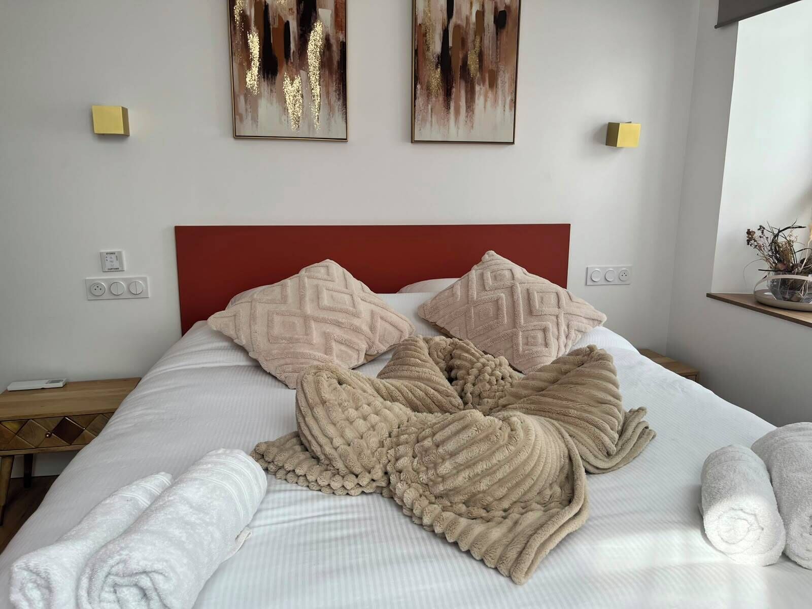 Ein ordentlich bezogenes Bett mit rotem Kopfteil, zwei an der Wand montierte Lichtschalter und zwei abstrakte Gemälde darüber.