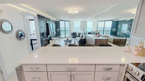 A modern kitchen with a central island, a dining area with a sofa and chairs, and a view through large windows. - Sundestin 1014 - Beautiful remodeled 1 BR sleeps 4 (Destin)