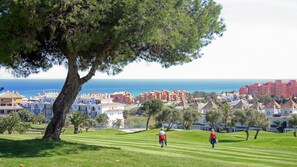 A coastal view with a tree-lined grassy area, a golf course, and a backdrop of colorful buildings and the sea. - Coto Real apartment Zira 321 (Manilva)