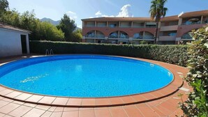 Una piscina rotonda con bordo piastrellato, circondata da un siepe e un edificio con balconi.