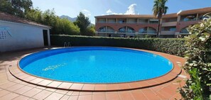 Ein runder Swimmingpool mit Fliesenplattform, umgeben von einem Hecken und einem Gebäude mit Balkonen.