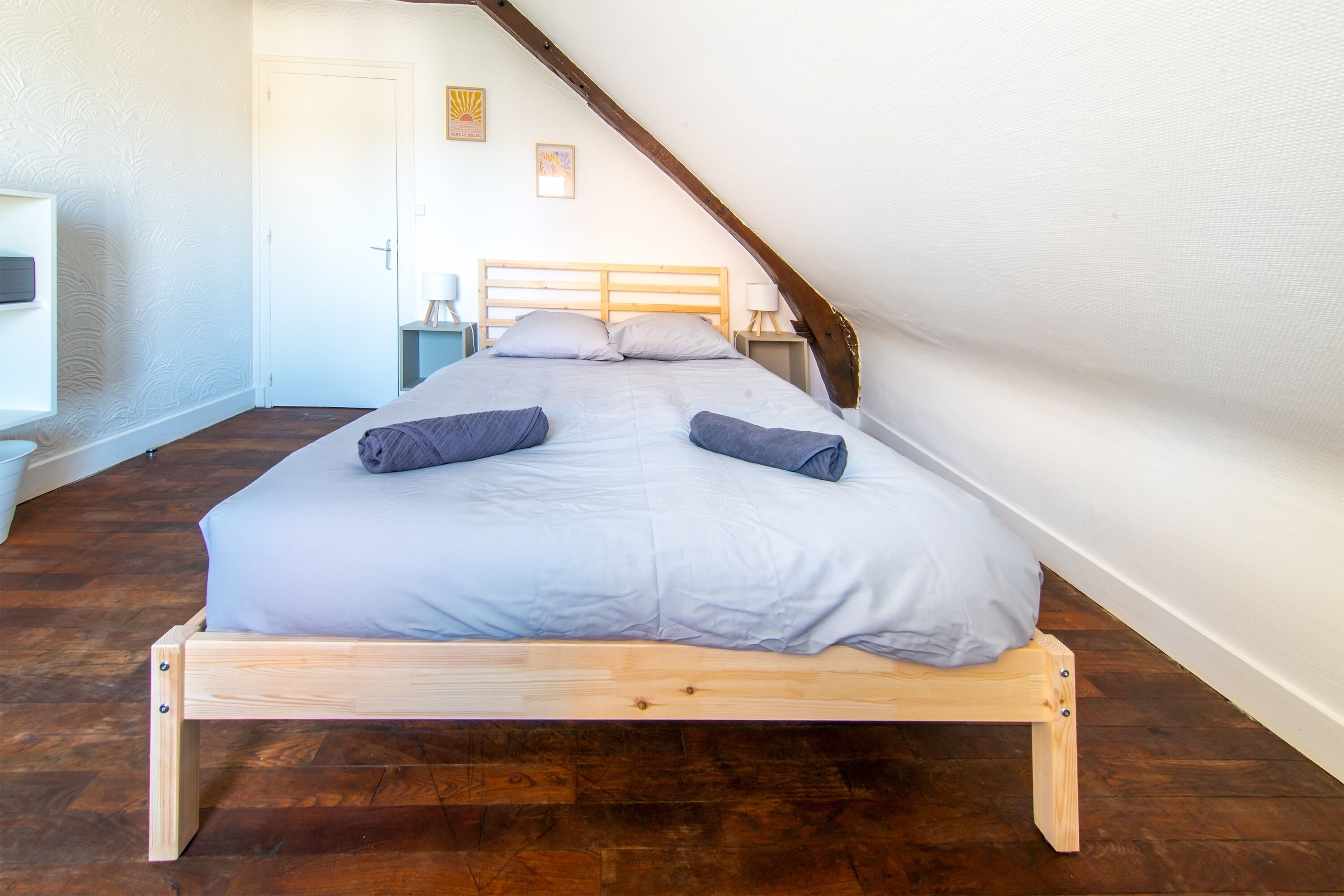 Un lit simple avec un cadre en bois et un matelas de couleur claire, dans une pièce dont le sol est recouvert de planches de bois.