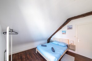 Une chambre avec un plafond en pente, un lit avec une literie bleue, une tête de lit en bois, une table de chevet avec une lampe et une porte.