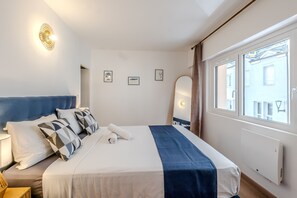 A hotel room with a bed, a nightstand, a lamp, a window with a view of buildings, and wall-mounted artwork. - La Yaute - Bel appt rénové à 2 pas du lac Léman (Evian-les-Bains)