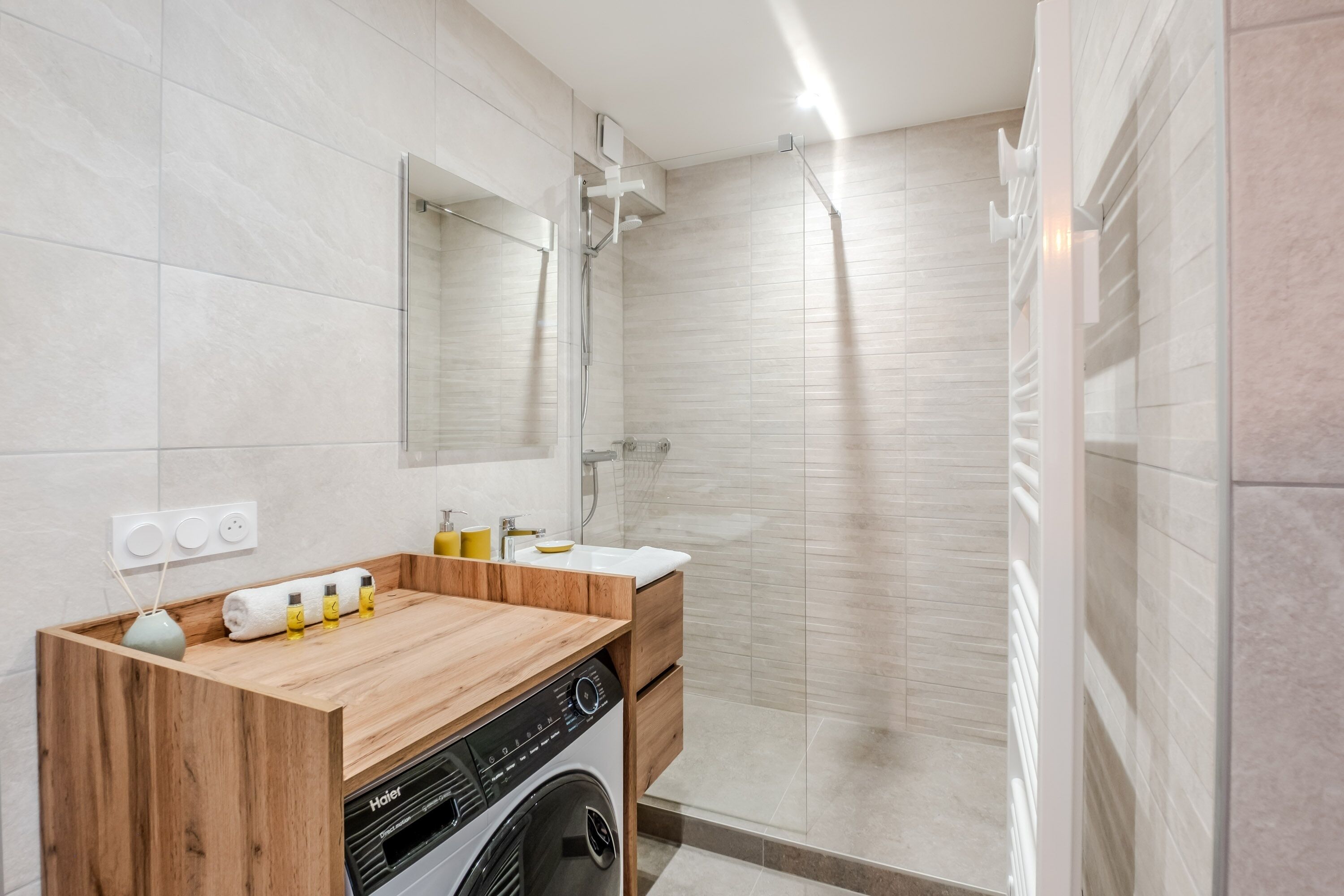A modern bathroom with a wooden vanity, a built-in washing machine, a glass shower enclosure, and a large mirror.