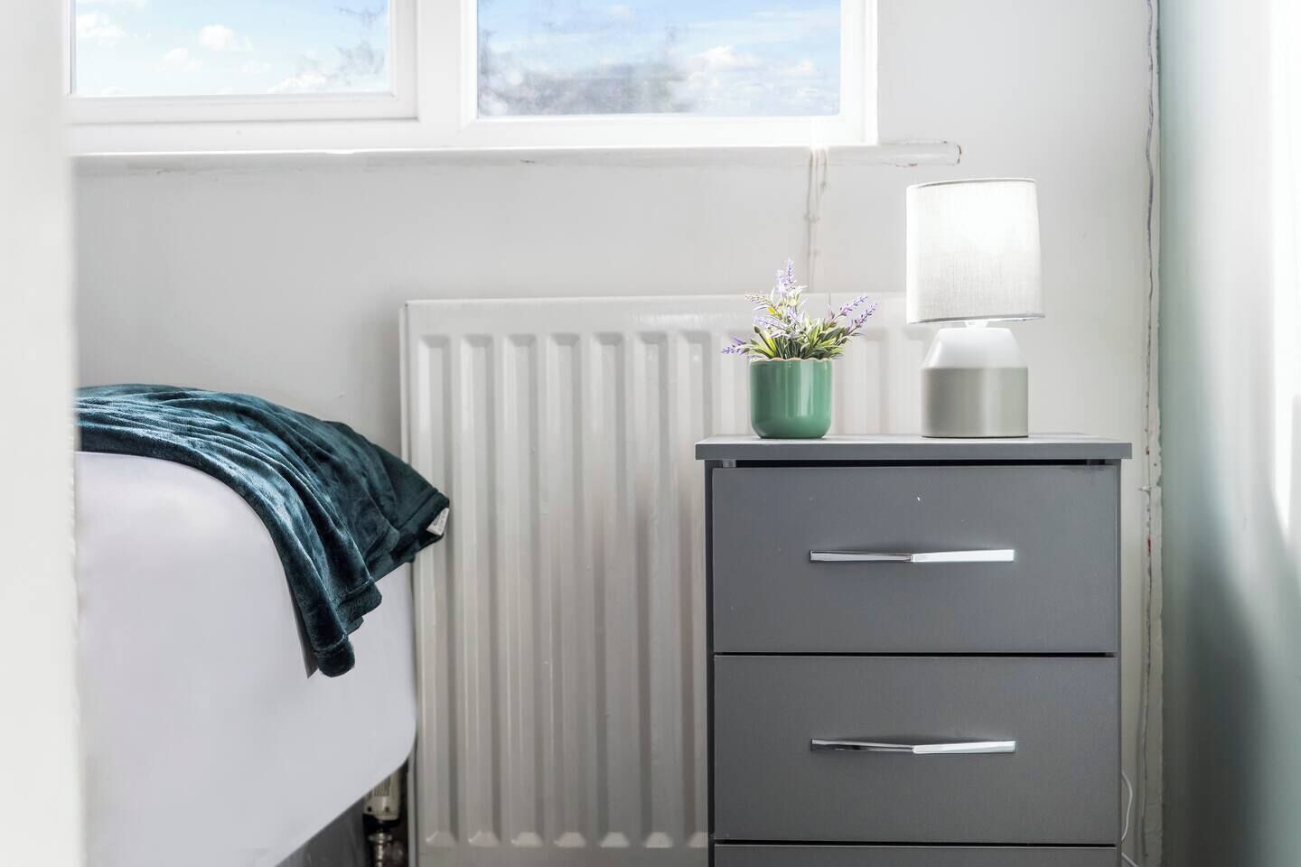 Un dormitorio con cama, radiador, escritorio con cajones, lámpara y ventana con vista.