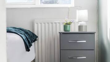 A bedroom with a bed, a radiator, a desk with drawers, a lamp, and a window with a view.